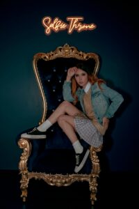 Fashionable young woman in denim jacket posing elegantly on a throne chair indoors.